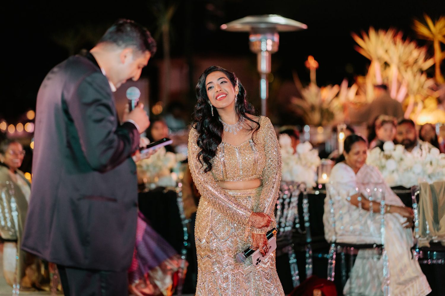 Indian bride in gold embroidered lehenga laughing joyfully as groom speaks into microphone at reception.