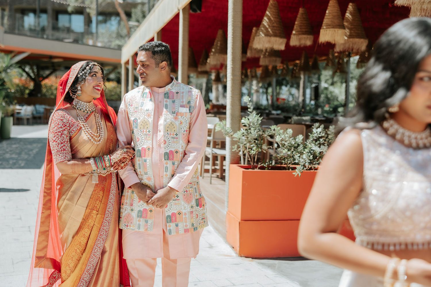 Indian bride in red saree and groom in white sherwani share a joyful moment at outdoor wedding venue.