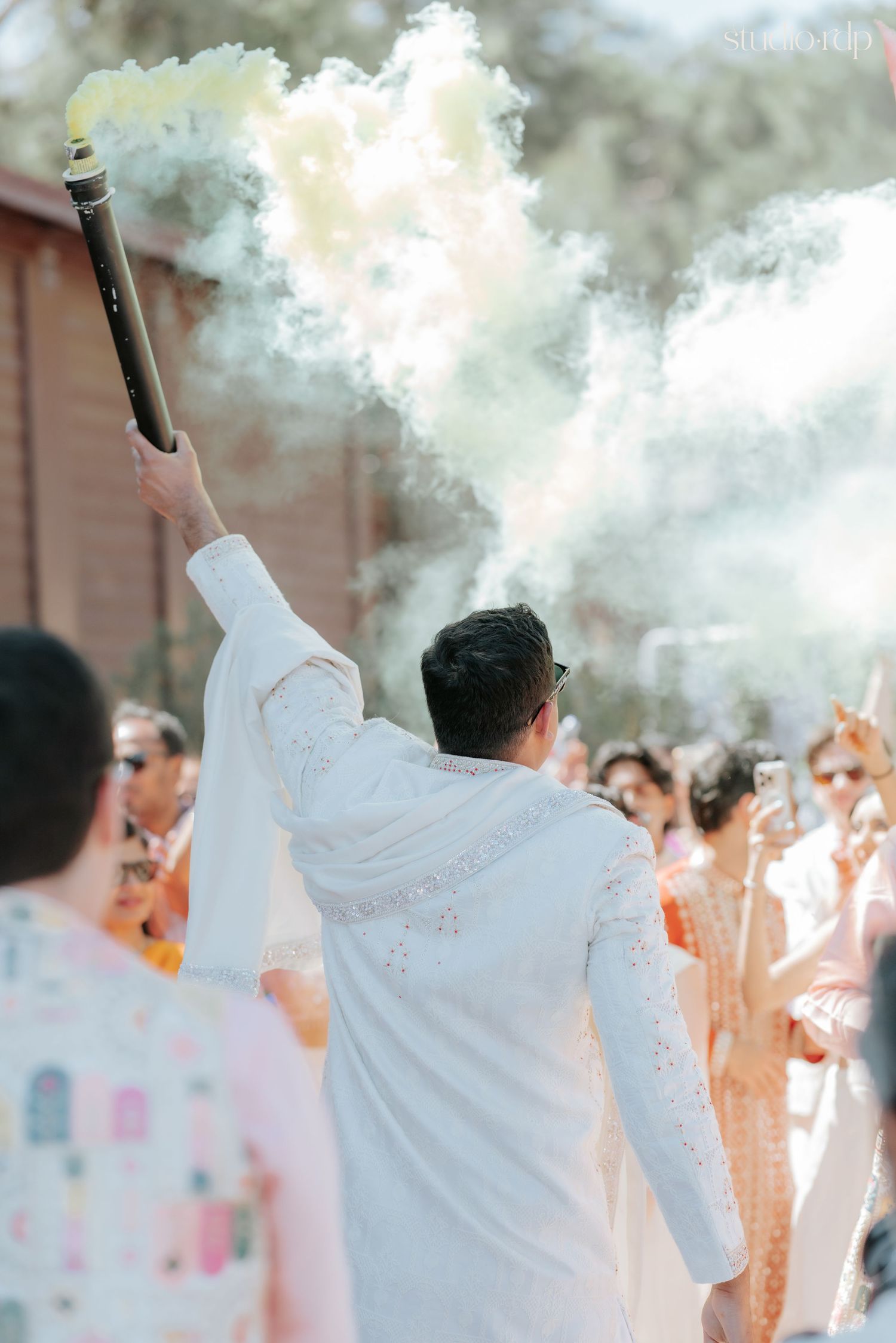 Groom in white sherwani holds smoke cannon at Indian wedding celebration outdoors.