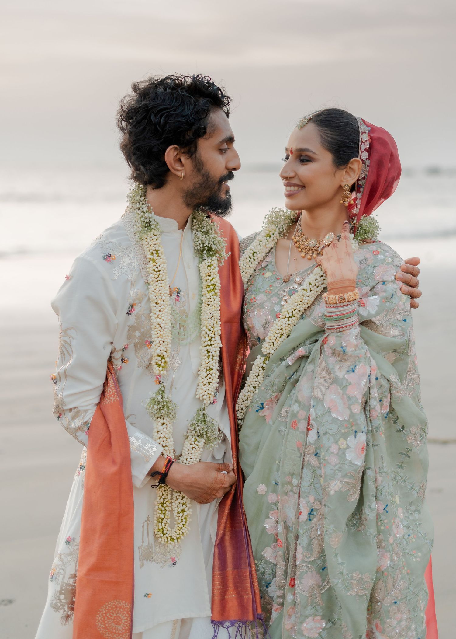 Couple in traditional Indian wedding attire with floral garlands posing on a beach at sunset.