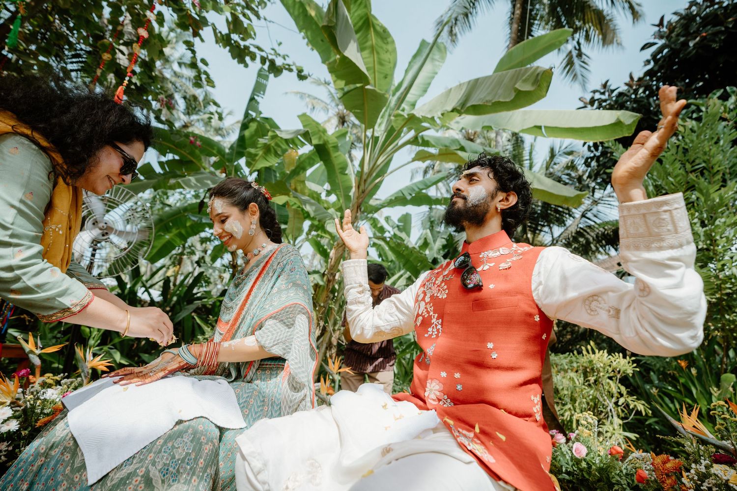 Couple in traditional Indian attire celebrate outdoors with colorful flowers and lush tropical greenery during a wedding ceremony.
