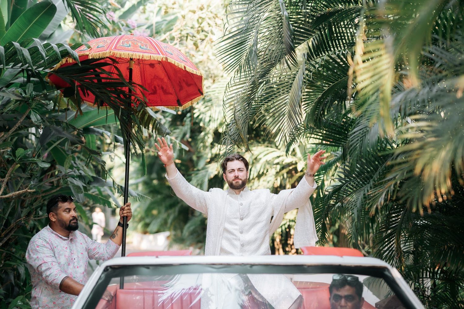 Groom in white kurta standing in car under red umbrella during baraat wedding procession.