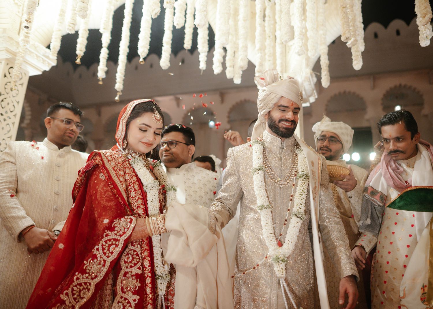 Indian bride in red lehenga and groom in white sherwani walk together at an elegant floral wedding ceremony.