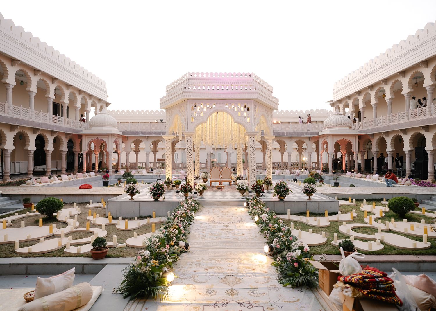 Ornate Indian wedding venue with white Mughal architecture, floral aisle, and manicured gardens in an open-air courtyard.
