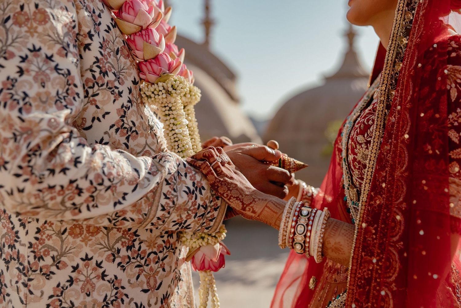 Hand adorned with traditional gold jewelry, bangles, and henna holds a red embroidered fabric with intricate patterns.