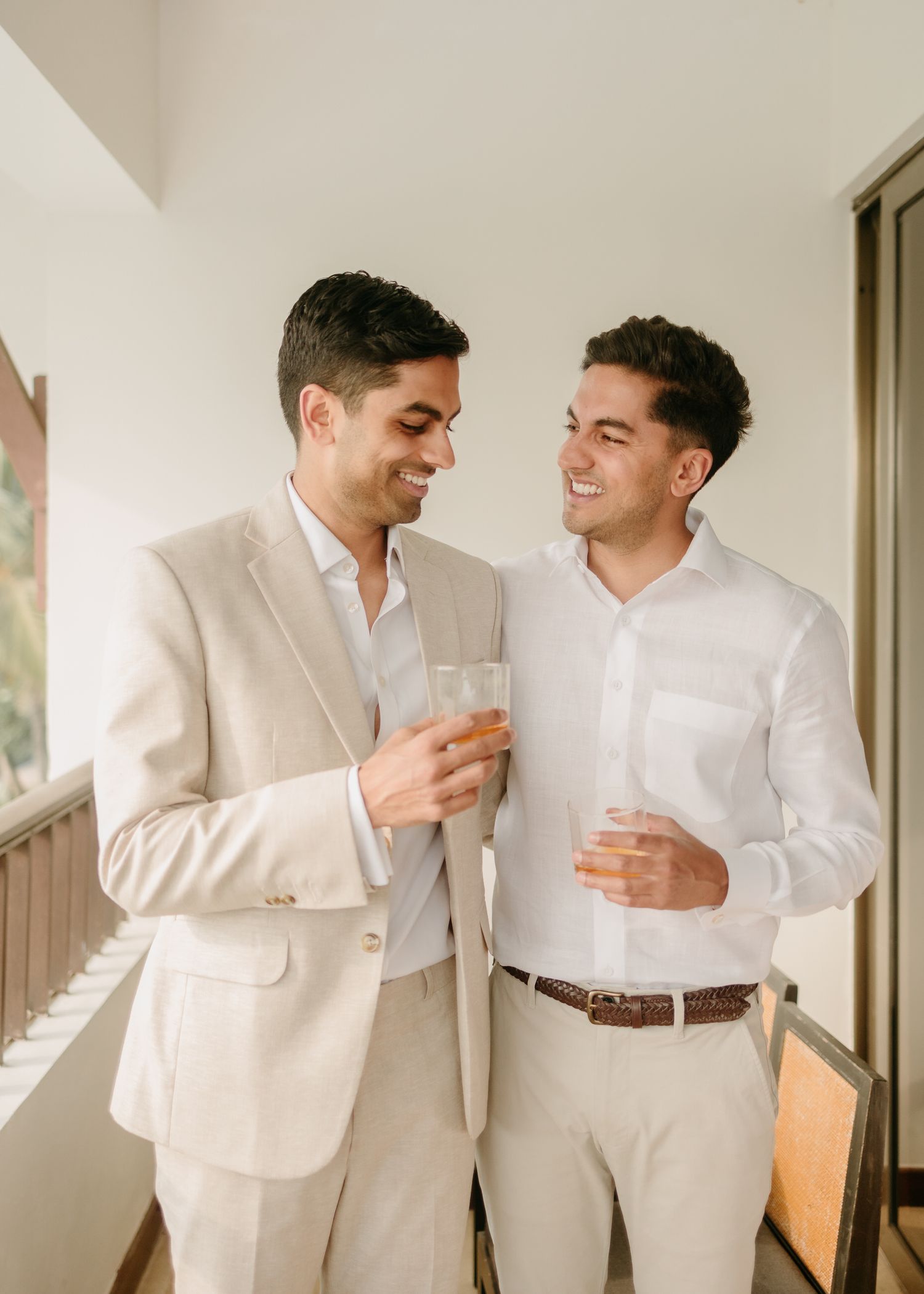 Two people in light colored attire share drinks and laughter together on a balcony.