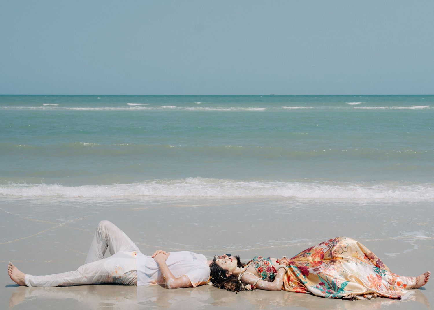 Two people in casual summer outfits lying down and relaxing on a sandy beach with turquoise ocean waves in the background.