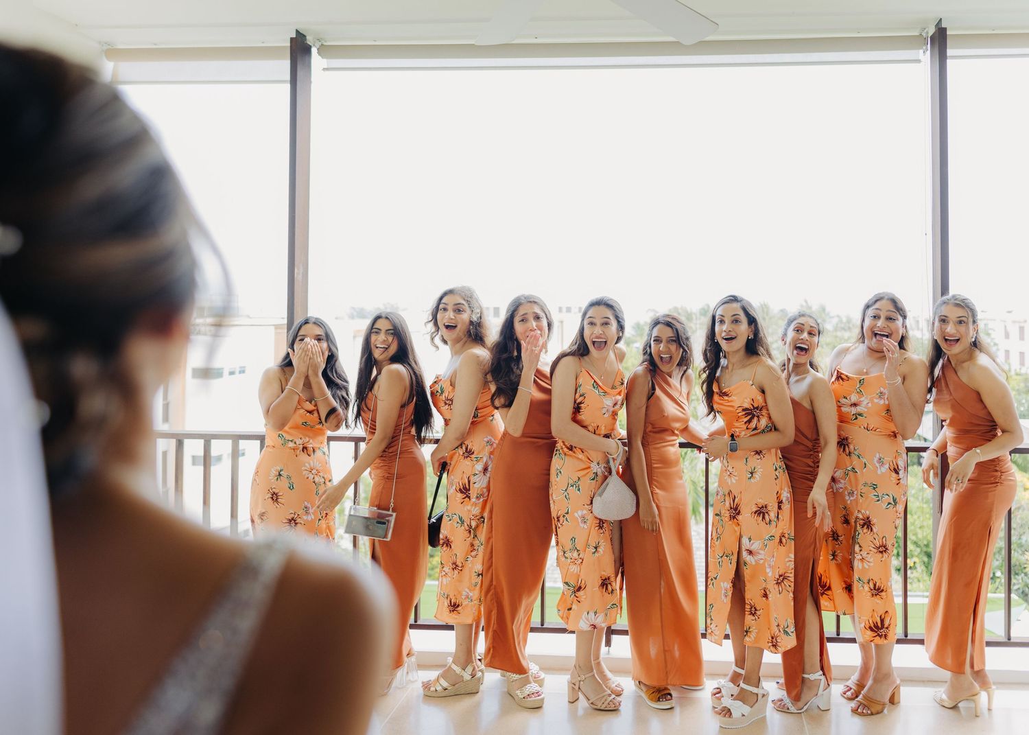 A group of people wearing elegant rust-colored bridesmaid dresses pose in a bright white room together.