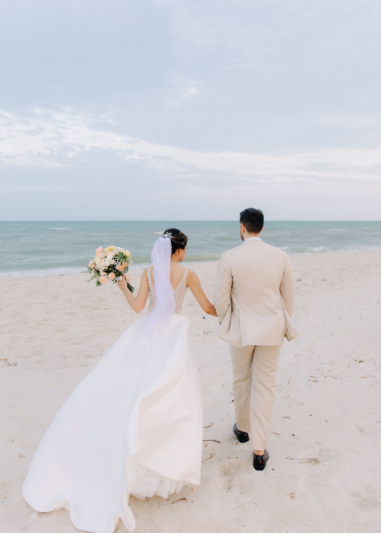 A couple walking hand in hand on a beach during their wedding, with the bride holding a white floral bouquet.