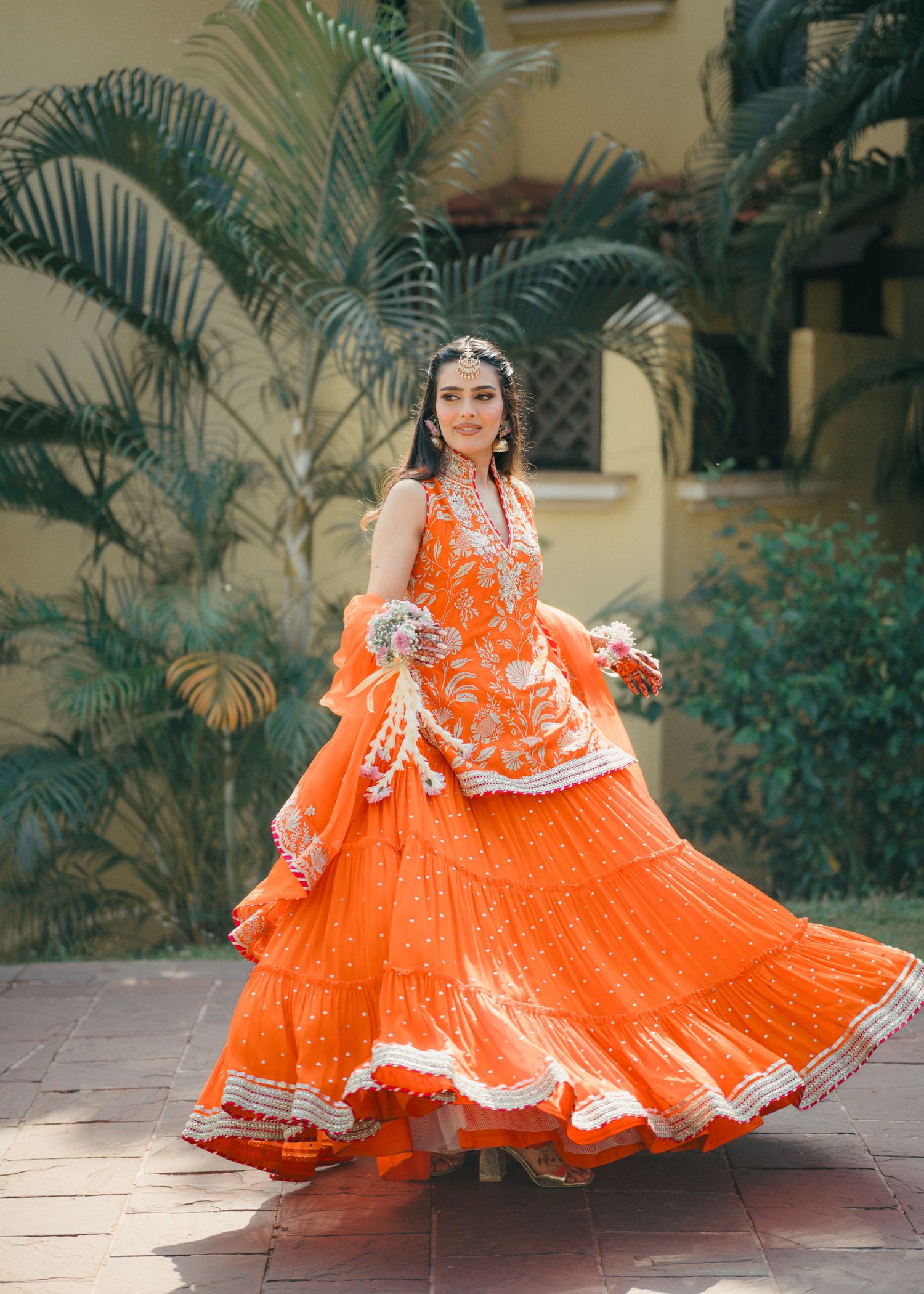 Person in vibrant orange and white traditional lehenga twirls against a tropical palm garden backdrop.