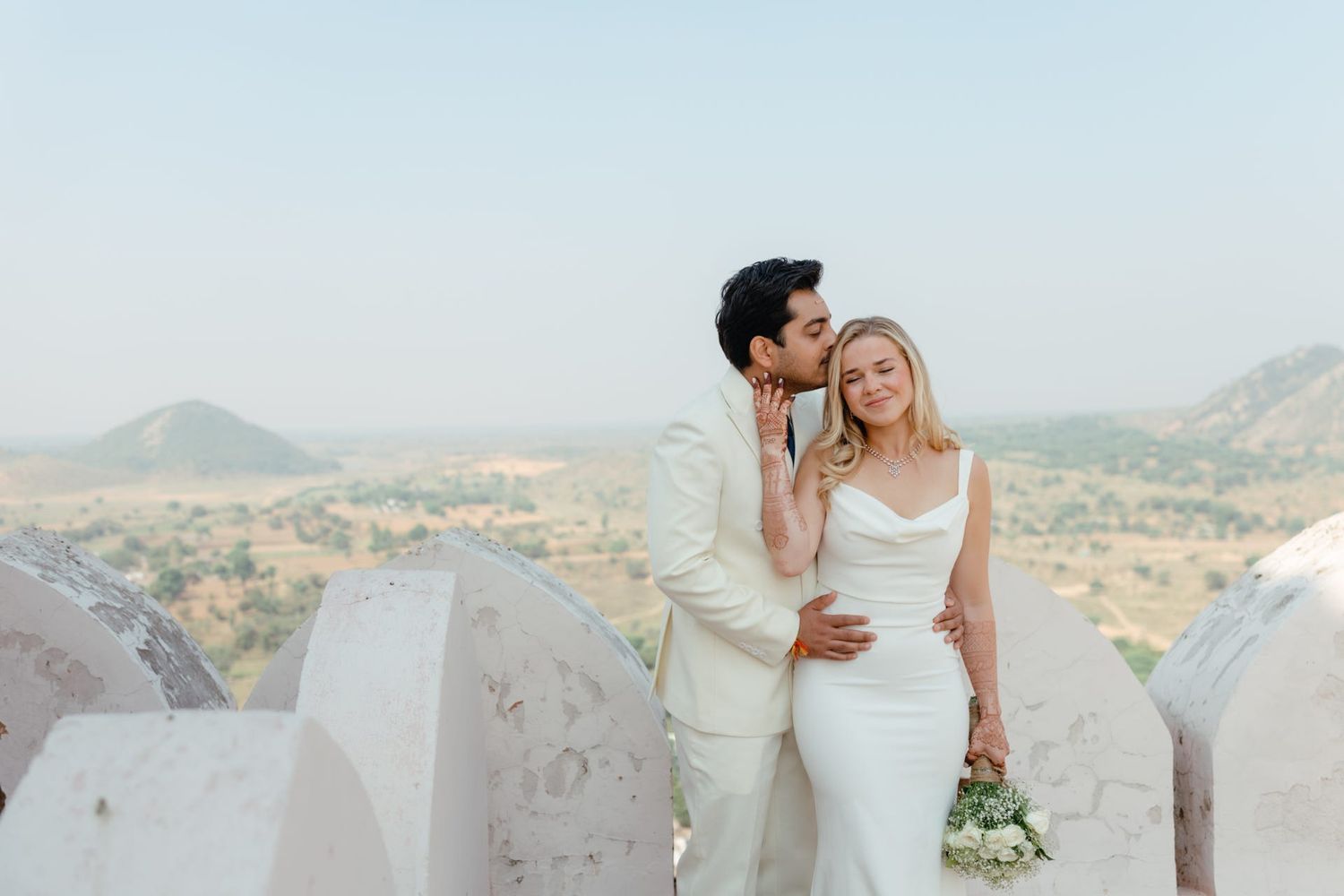 Couple in white attire embracing on rooftop with mountain landscape in background.