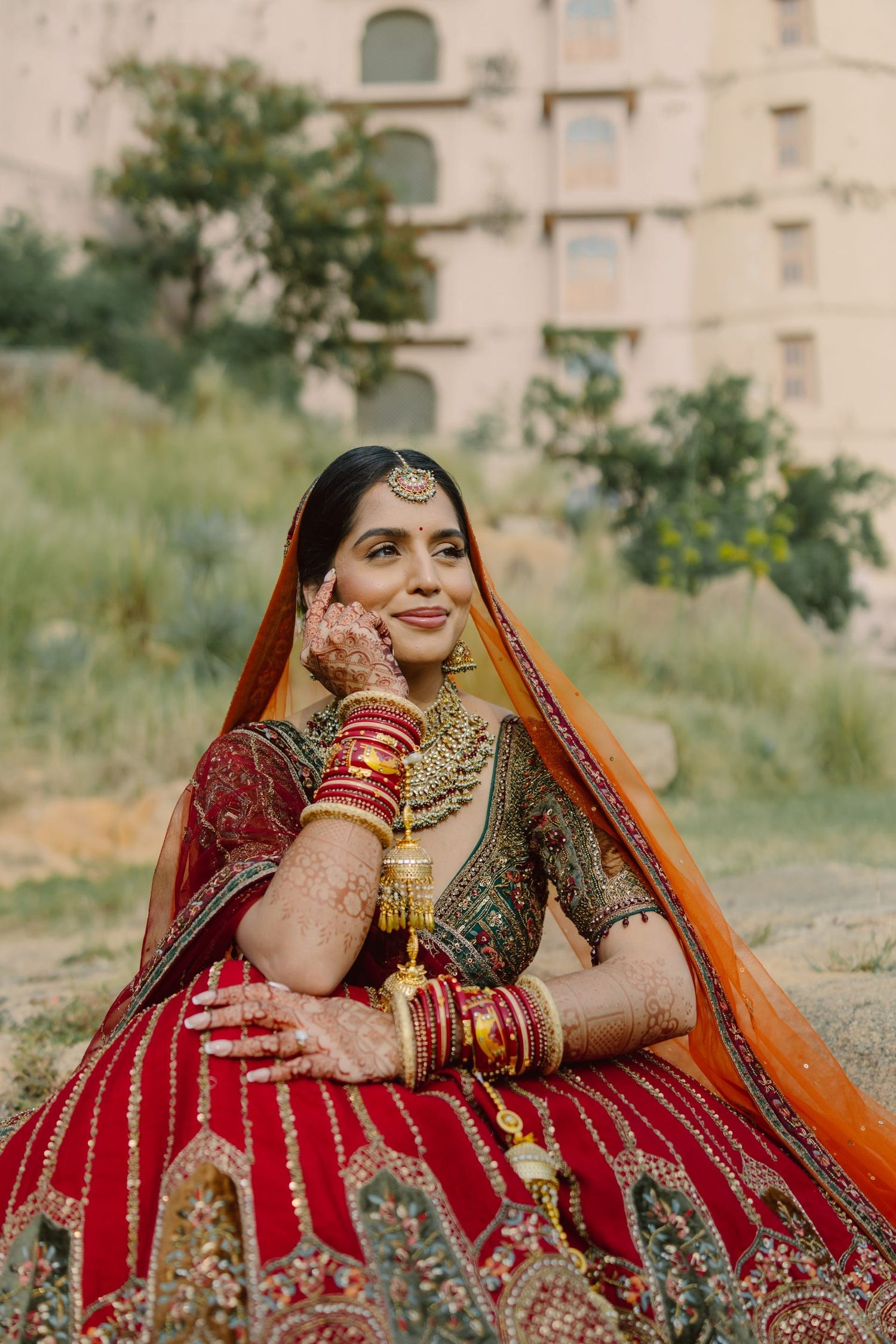 A bride in red and gold traditional attire poses against a building backdrop.