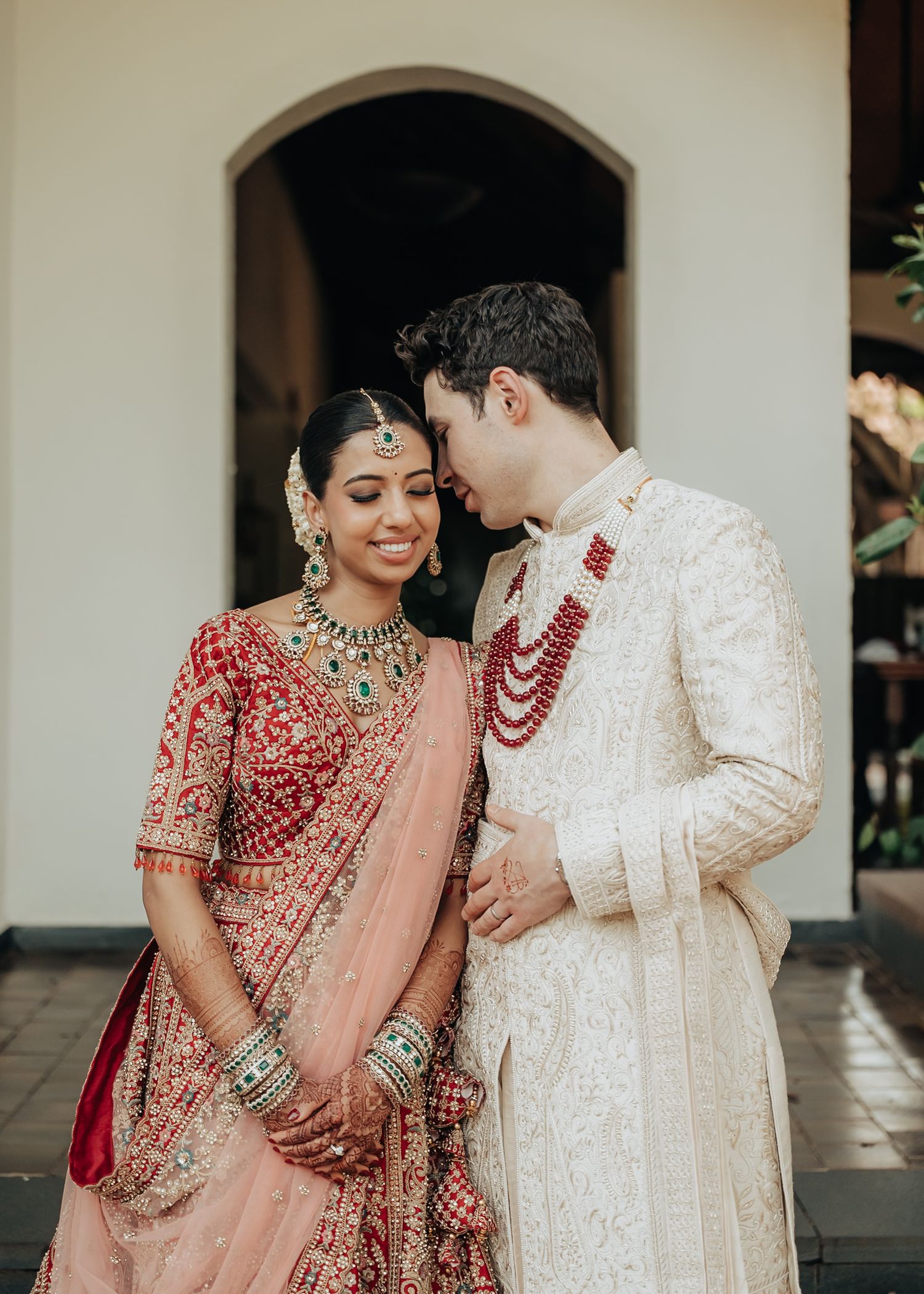 A couple in traditional Indian wedding attire shares an intimate moment in a covered walkway.
