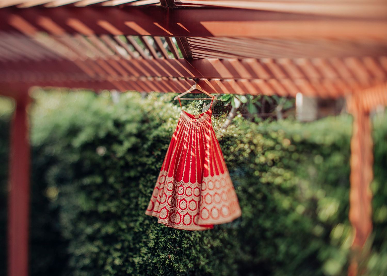 A traditional red and gold Indian bridal lehenga dress hangs from a wooden pergola in an outdoor garden setting.
