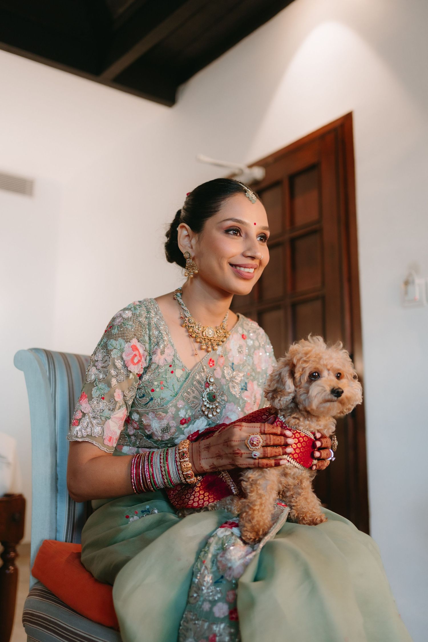 Indian bride in mint saree holding a fluffy dog, wearing gold jewelry and bangles, smiling joyfully indoors.