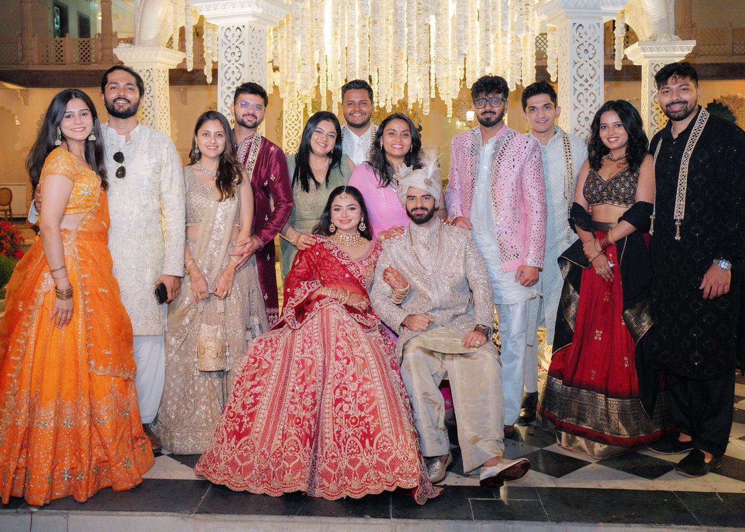 Indian wedding group photo with bride in red lehenga and groom in white sherwani surrounded by family and friends.