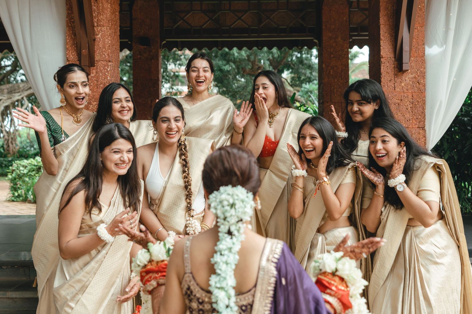 Bridesmaids in cream gold sarees joyfully react to bride during traditional Indian wedding ceremony.