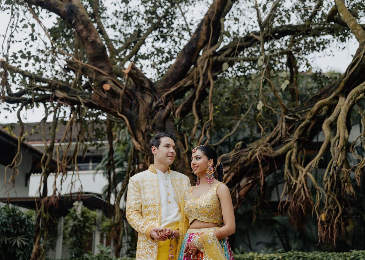 Couple in traditional Indian wedding attire posing before a grand banyan tree, groom in gold sherwani, bride in yellow lehenga.