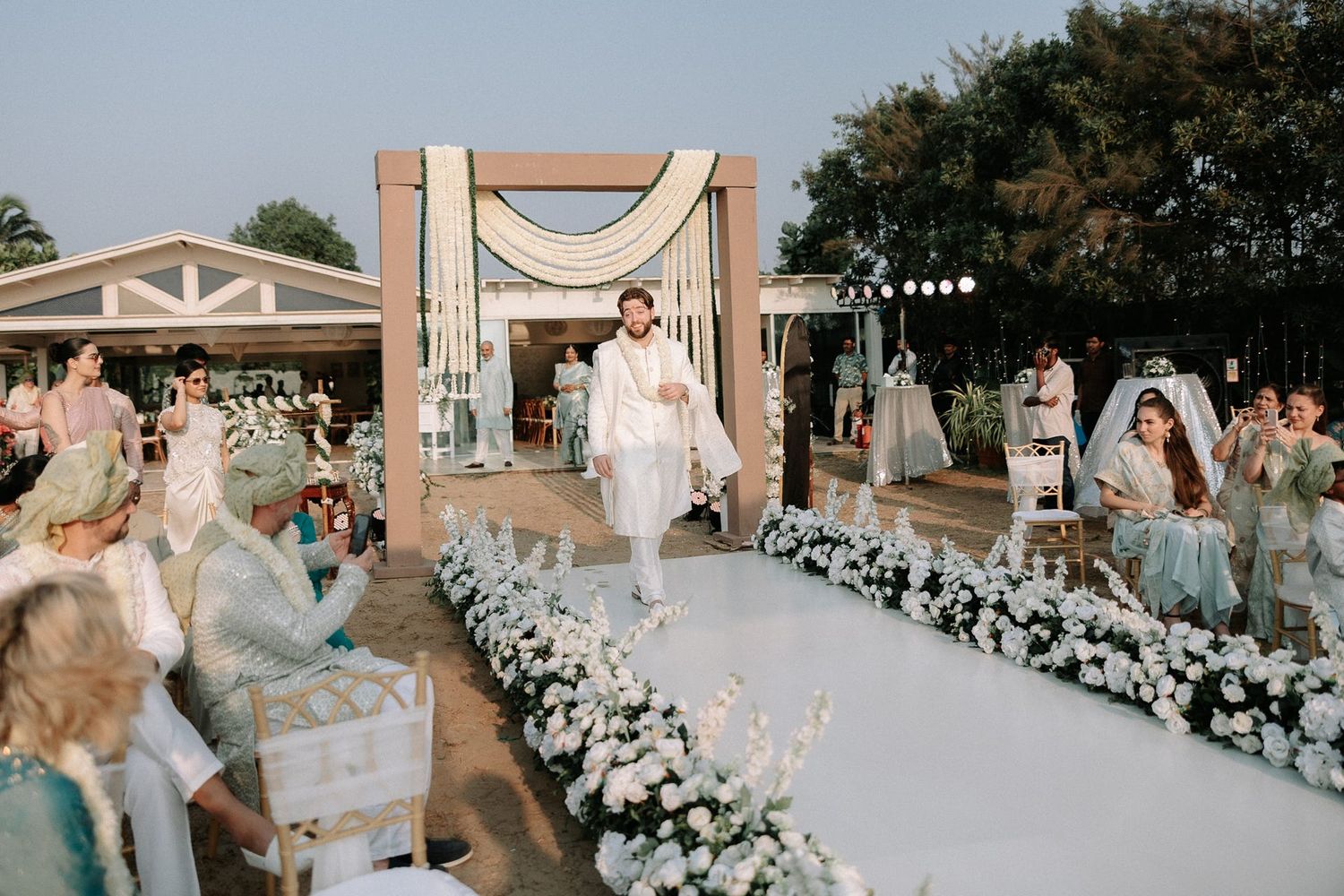 Groom walks down floral-lined white aisle at elegant outdoor South Asian wedding ceremony with guests seated.