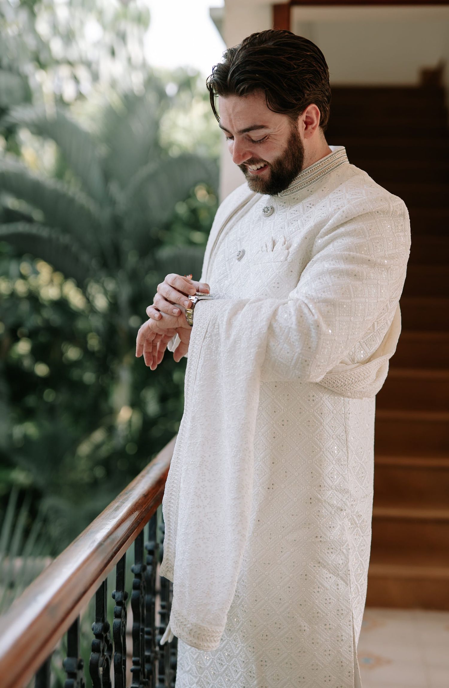 A groom in an embroidered white sherwani adjusts his cufflinks on a balcony surrounded by lush greenery.