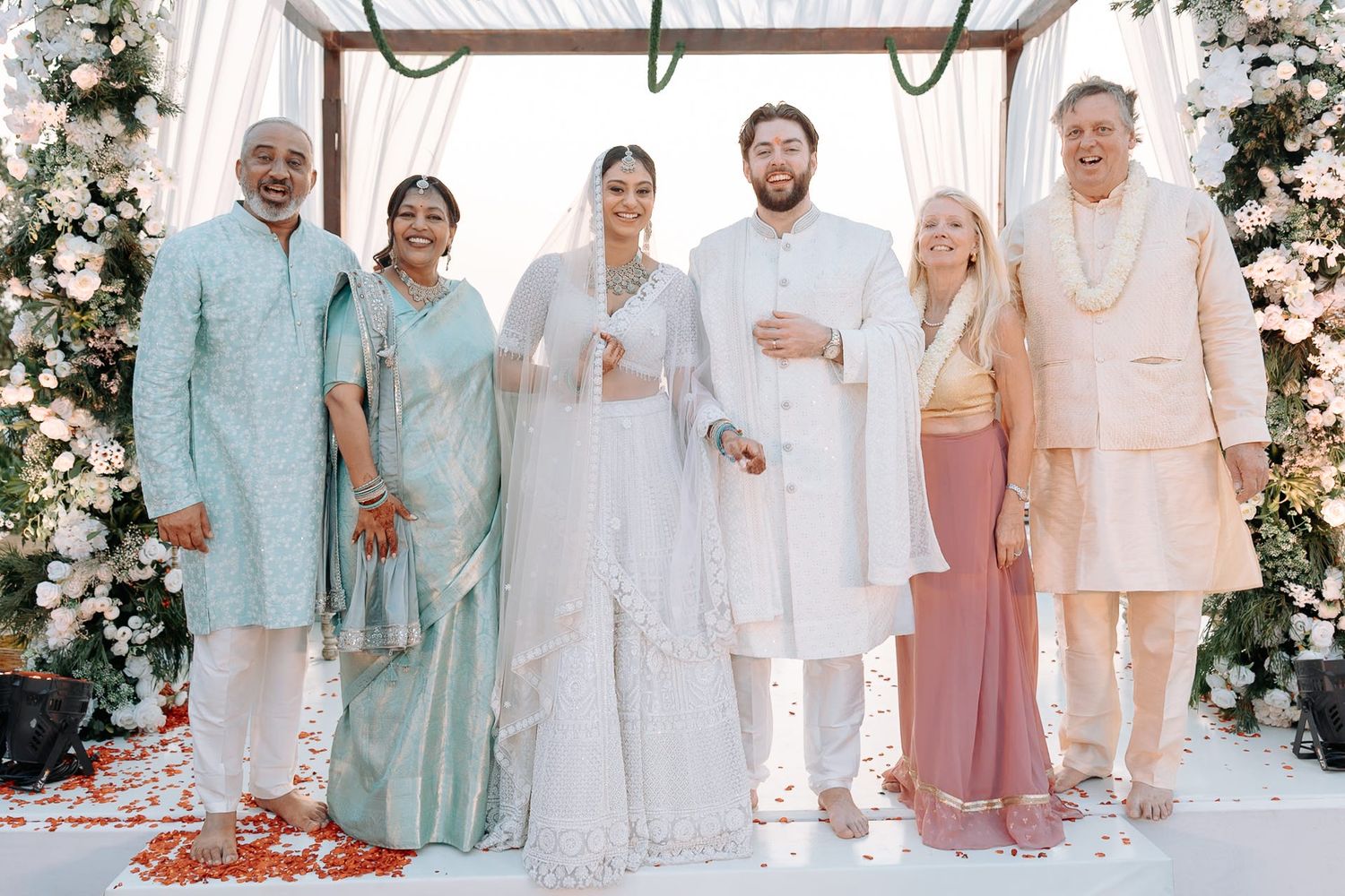 Six guests in elegant Indian and Western attire pose together at a floral wedding ceremony backdrop.