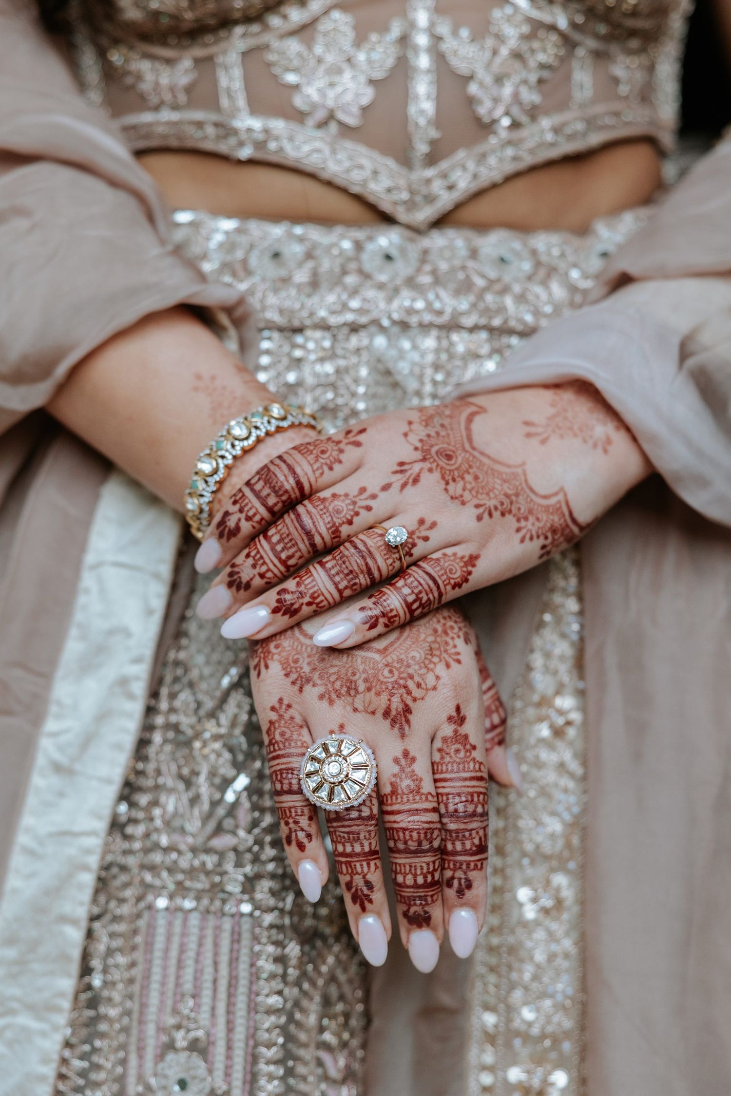 Bride's hands adorned with intricate henna mehndi designs, diamond rings, and gold bracelet against embroidered lehenga.
