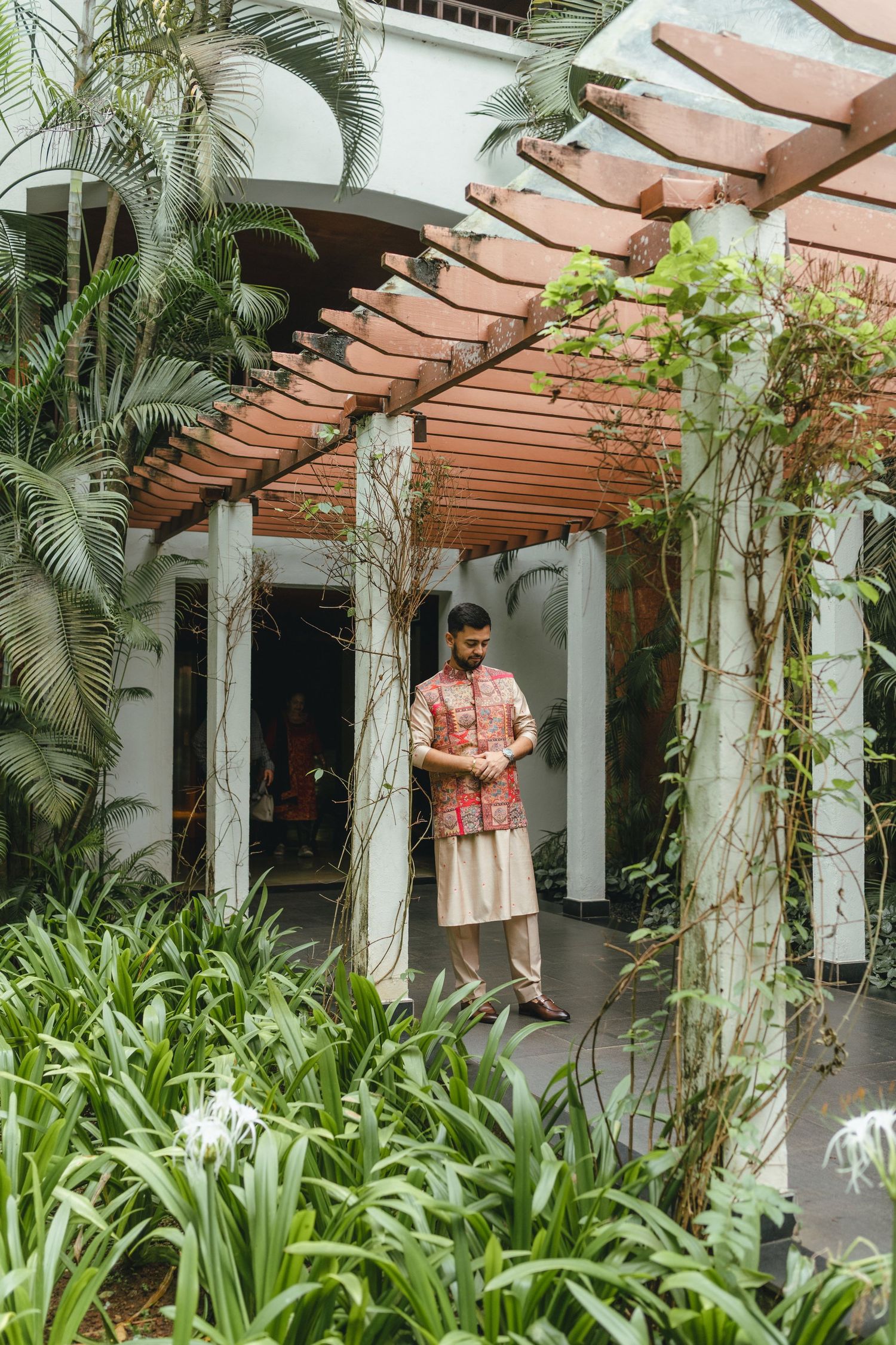 Man in traditional Indian kurta stands under wooden pergola surrounded by lush tropical greenery.