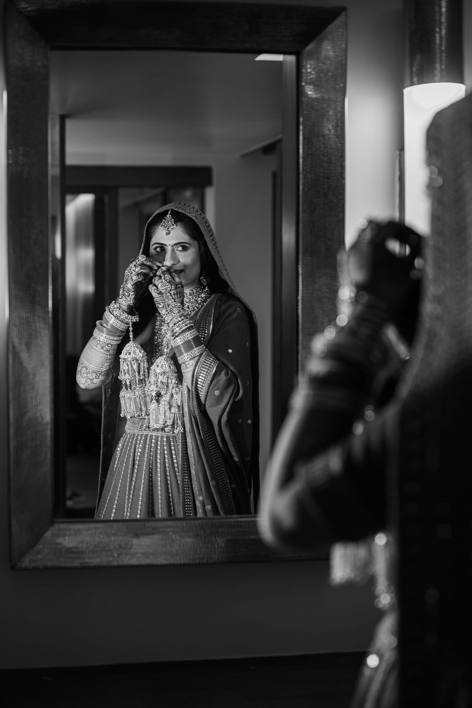 Black and white photo of an Indian bride in traditional bridal attire adjusting her veil, reflected in a ornate mirror.