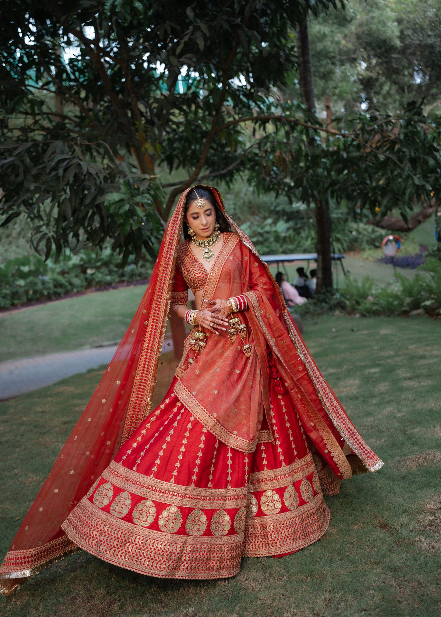 Bride in red and gold embroidered lehenga with dupatta twirling in garden setting.