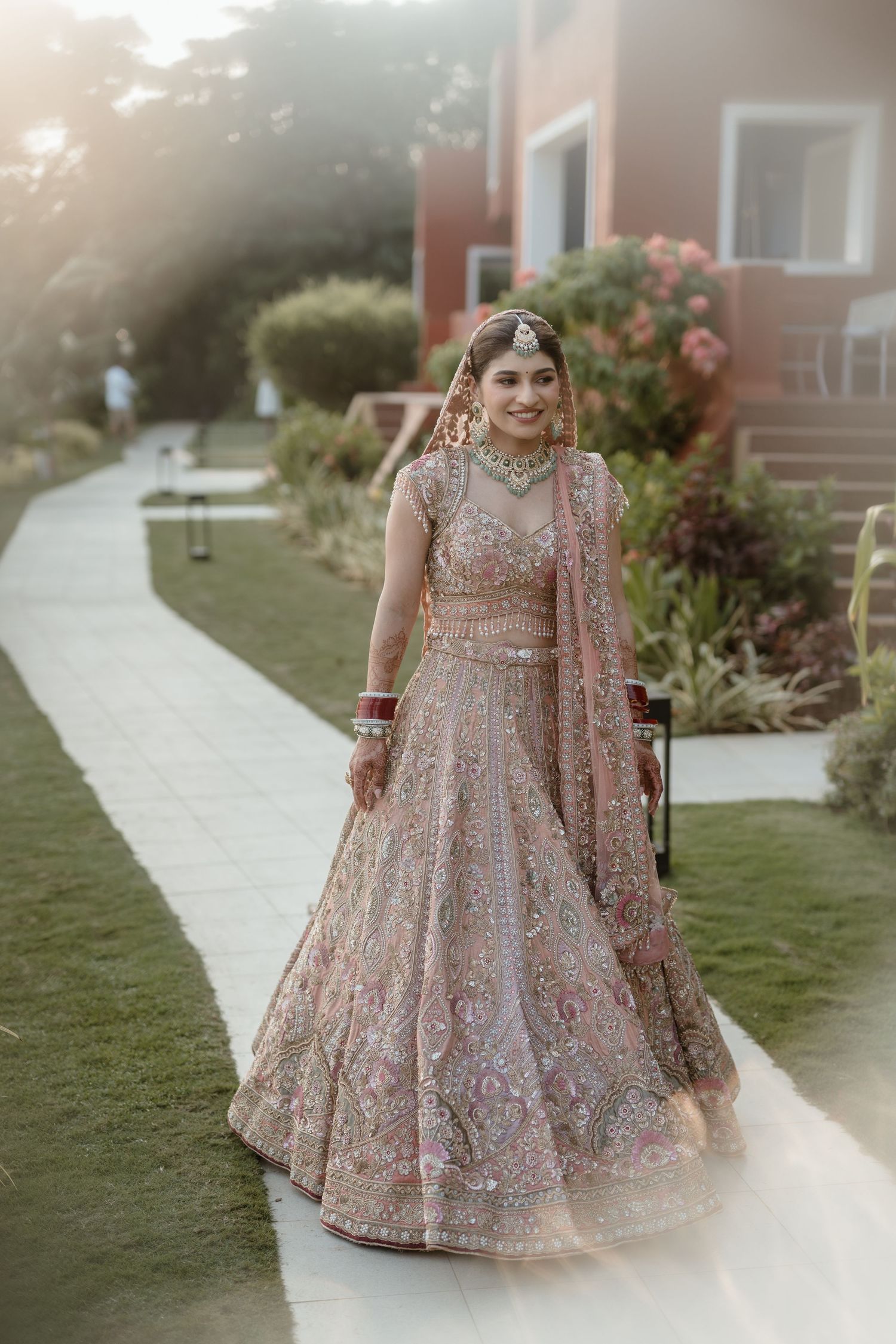 A person wearing a shimmering pink and gold embellished Indian lehenga poses in a garden setting at sunset.