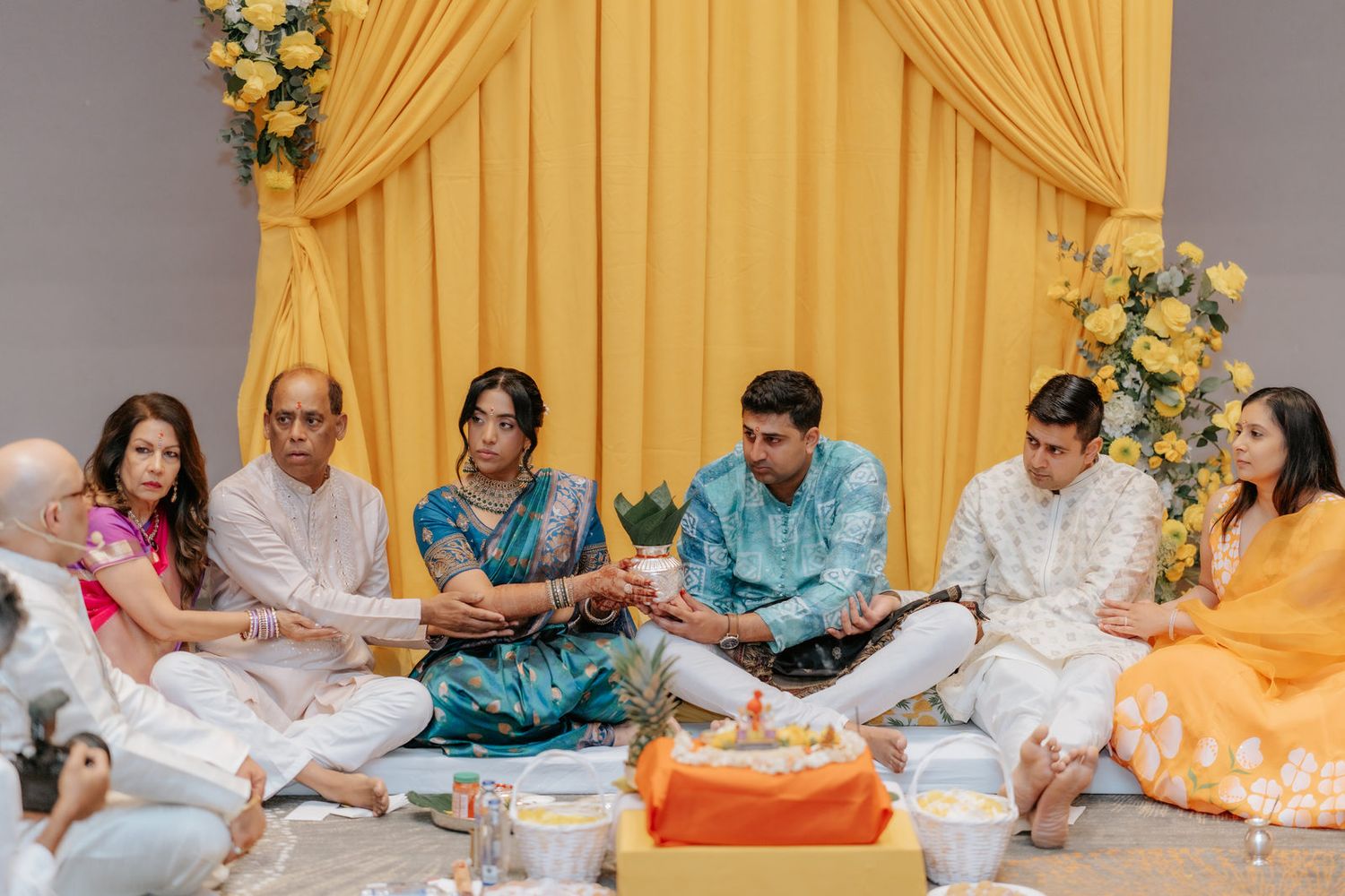 Indian family gathered for traditional Hindu puja ceremony, seated on floor before yellow draped altar with flowers.