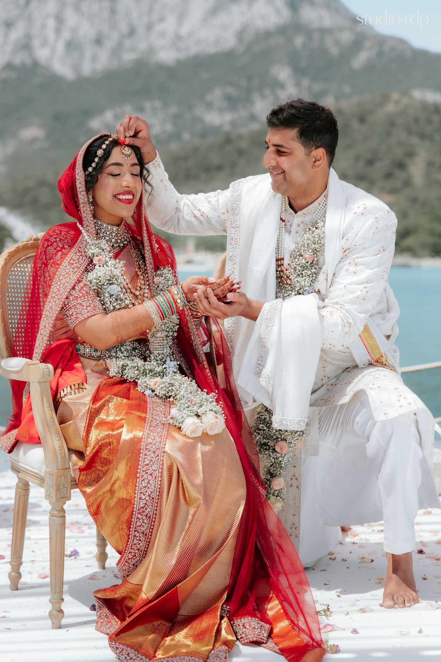 Bride in red saree and groom in white sherwani perform Hindu wedding rituals outdoors with mountain backdrop.