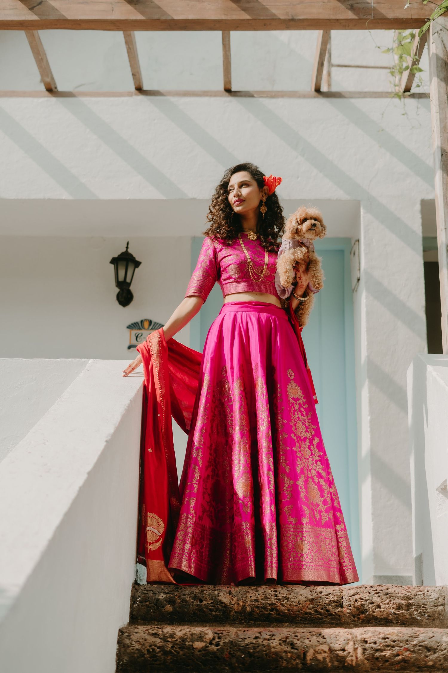 Woman in pink lehenga holding a small dog on steps of a white building.
