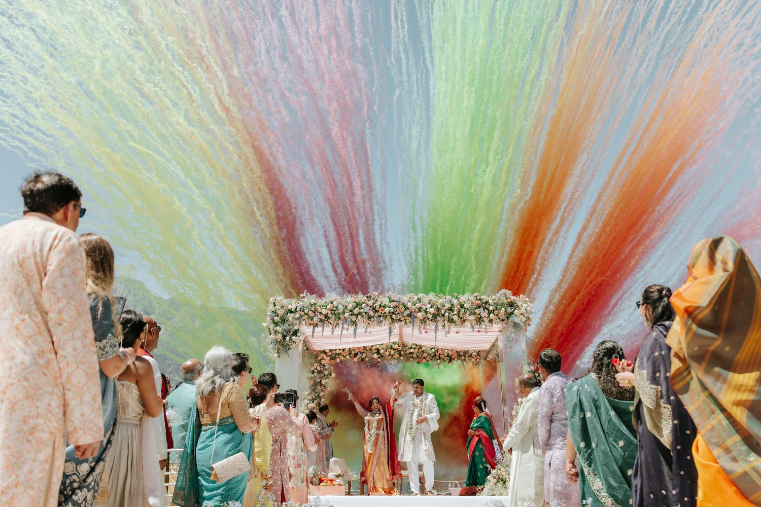 Colorful streamers cascade over an Indian wedding ceremony with guests in traditional attire gathered around a floral mandap.