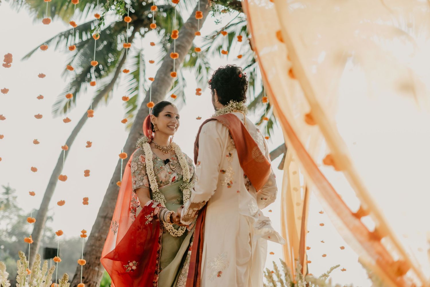 Indian bride and groom in traditional attire holding hands under floral decorations at an outdoor wedding ceremony.