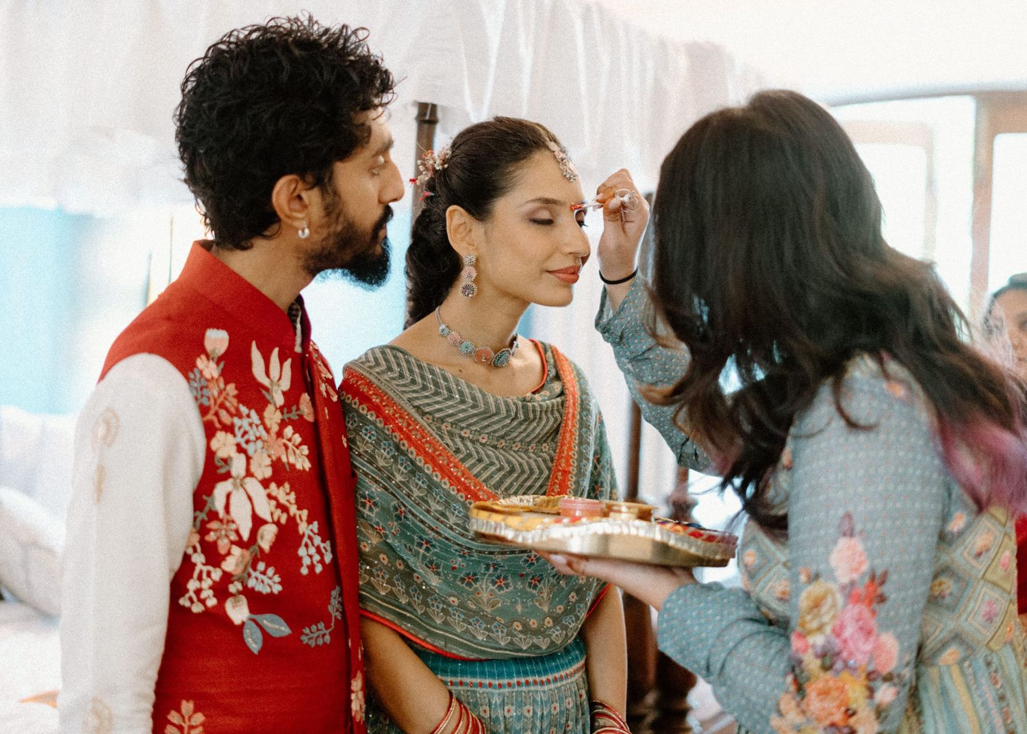 A woman applies makeup to a bride in traditional Indian attire, while a groom in red embroidered sherwani stands beside her.