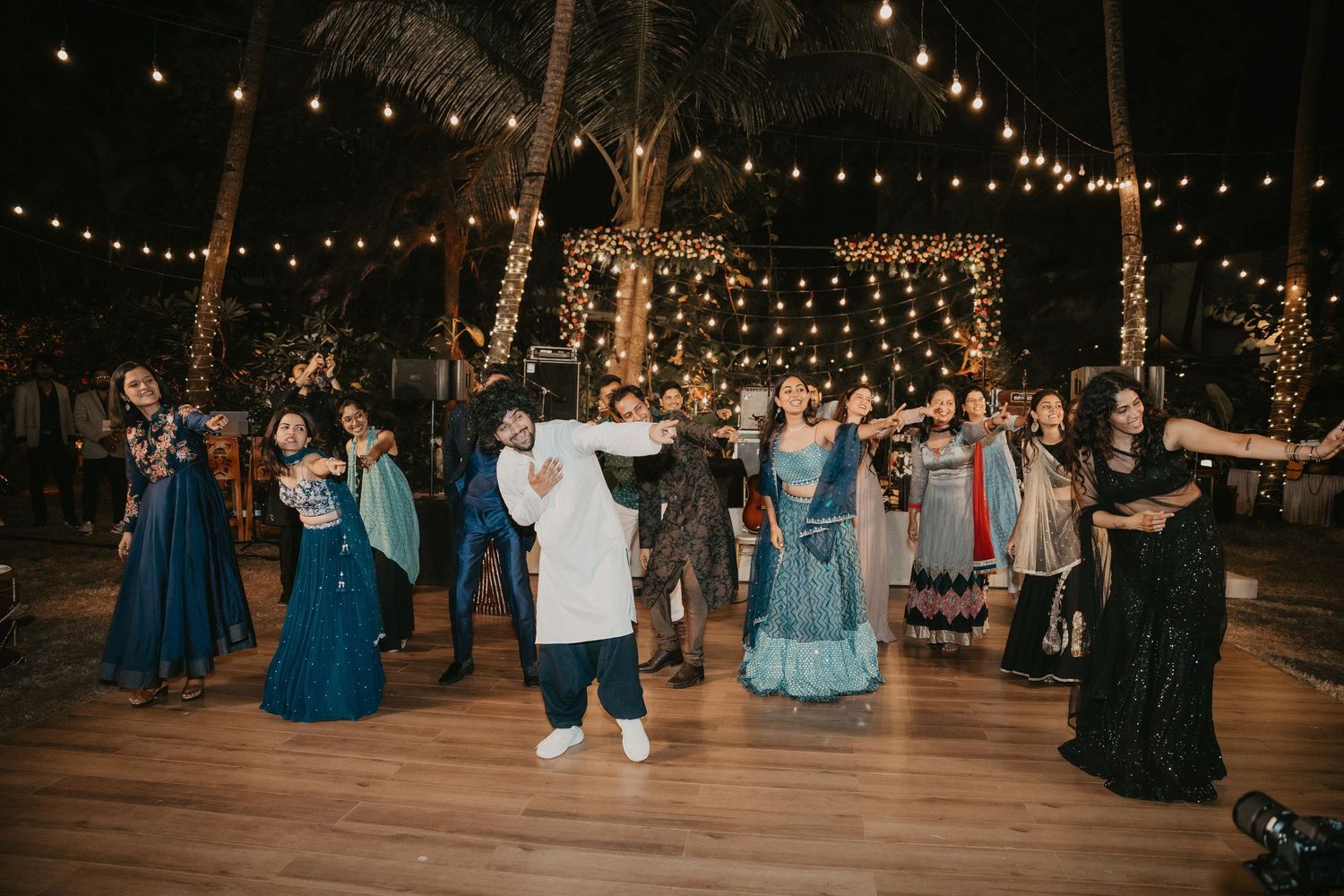 Group of guests dancing joyfully at an Indian wedding reception with string lights and palm trees.