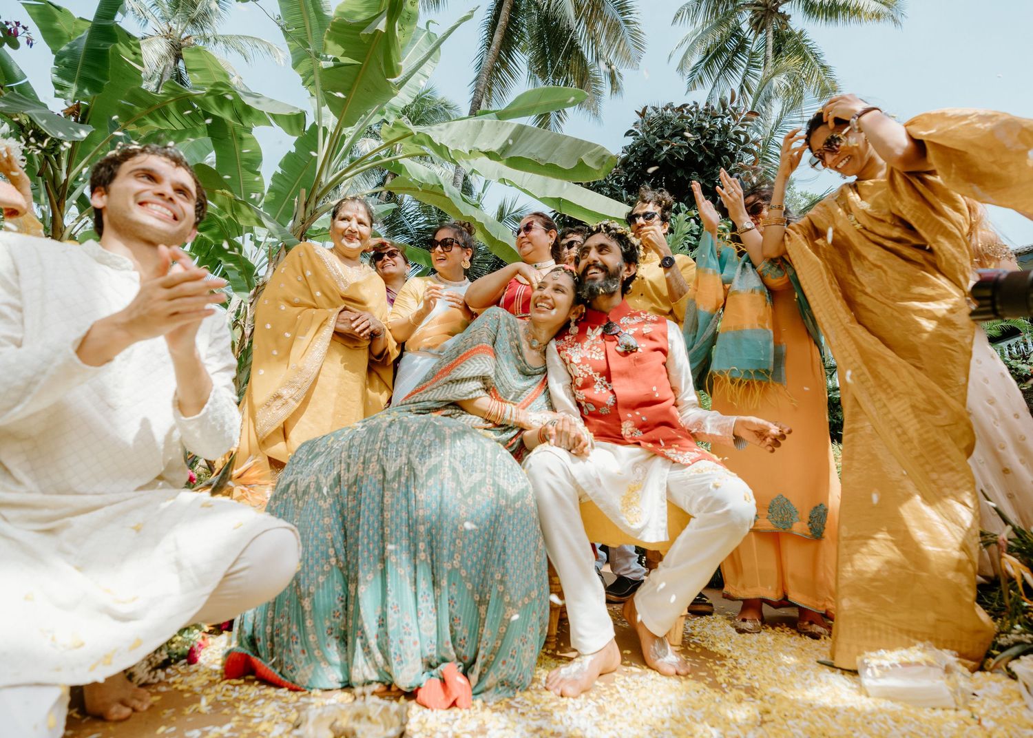Joyful Indian wedding celebration with guests in colorful traditional attire outdoors near tropical palm trees.