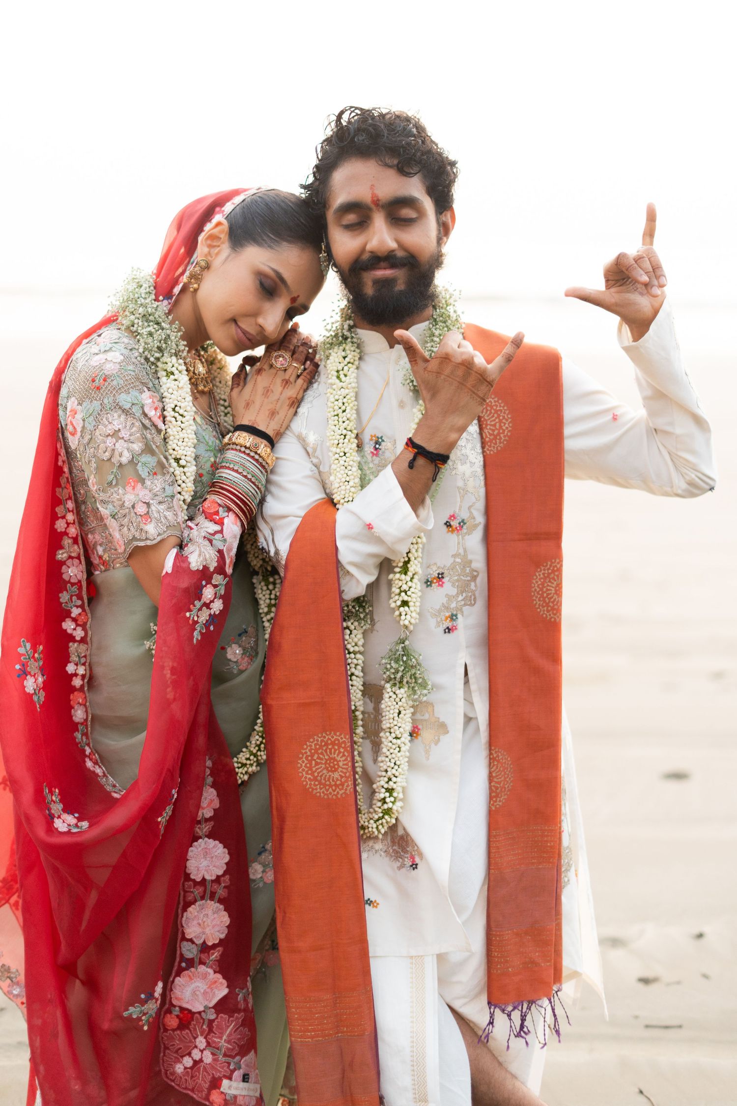 Indian couple in traditional wedding attire with floral garlands posing on a beach, bride in red dupatta.