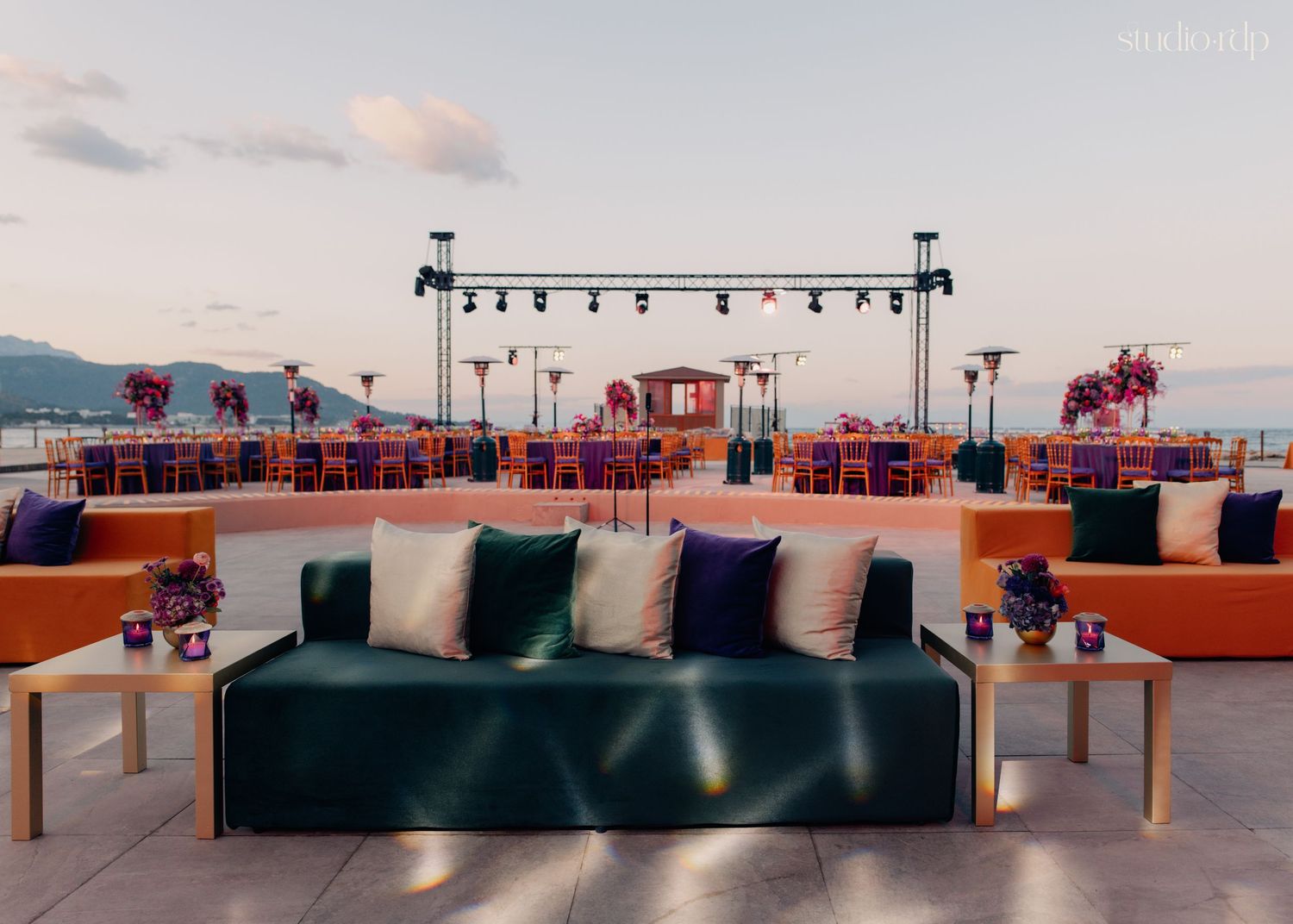 Elegant outdoor wedding venue with green velvet sofas, orange seating, and a decorated stage at sunset.