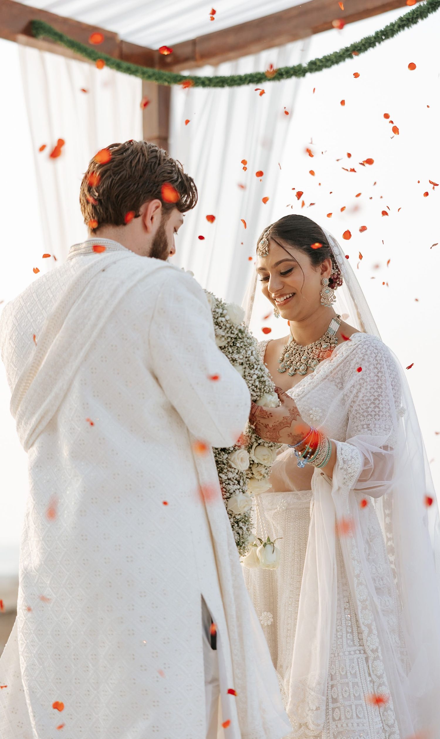 Couple in white traditional Indian wedding attire exchanging vows under floral arch with red petals falling.
