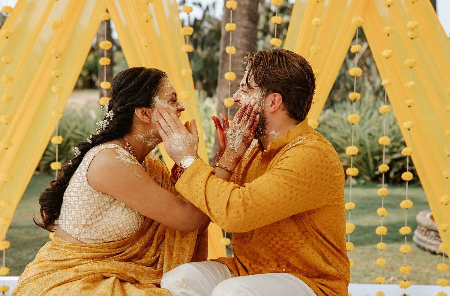 Couple in yellow Indian wedding attire playfully smearing haldi on each other's faces during a vibrant haldi ceremony.