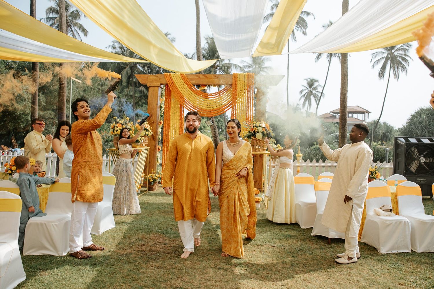Couple walks under yellow fabric canopy at vibrant Indian wedding ceremony outdoors with palm trees.