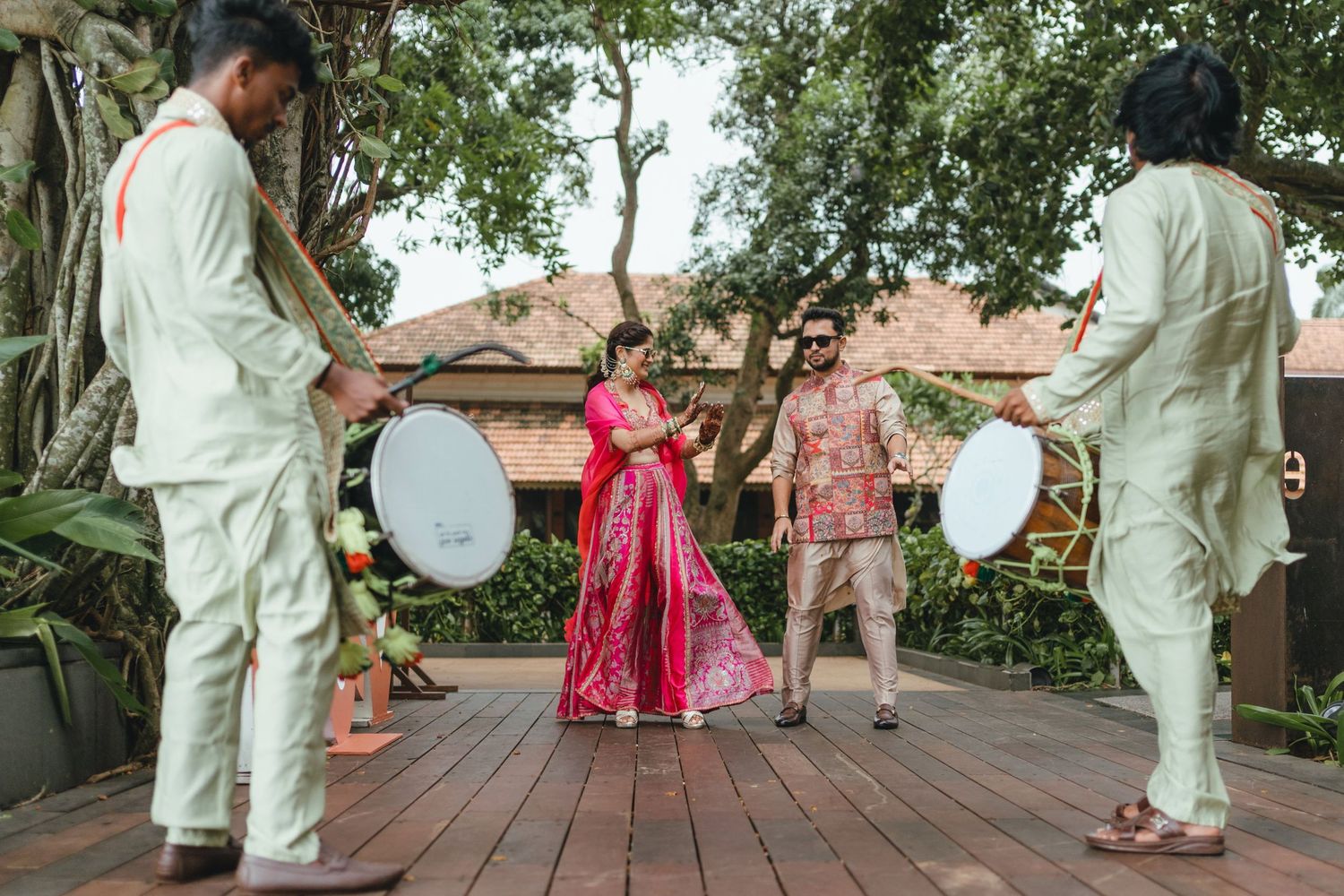 Bride in red lehenga with musicians playing dhol drums during Indian wedding celebration on wooden deck.