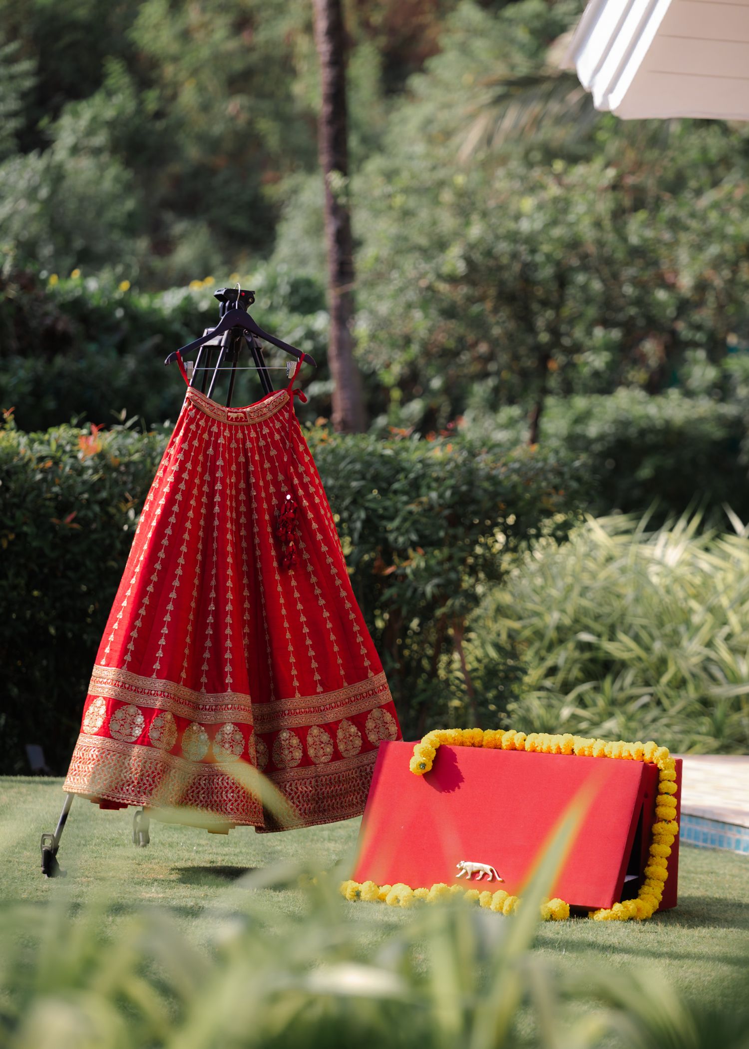 Red and gold traditional Indian lehenga displayed on hanger outdoors with matching red clutch adorned with yellow pom-poms.