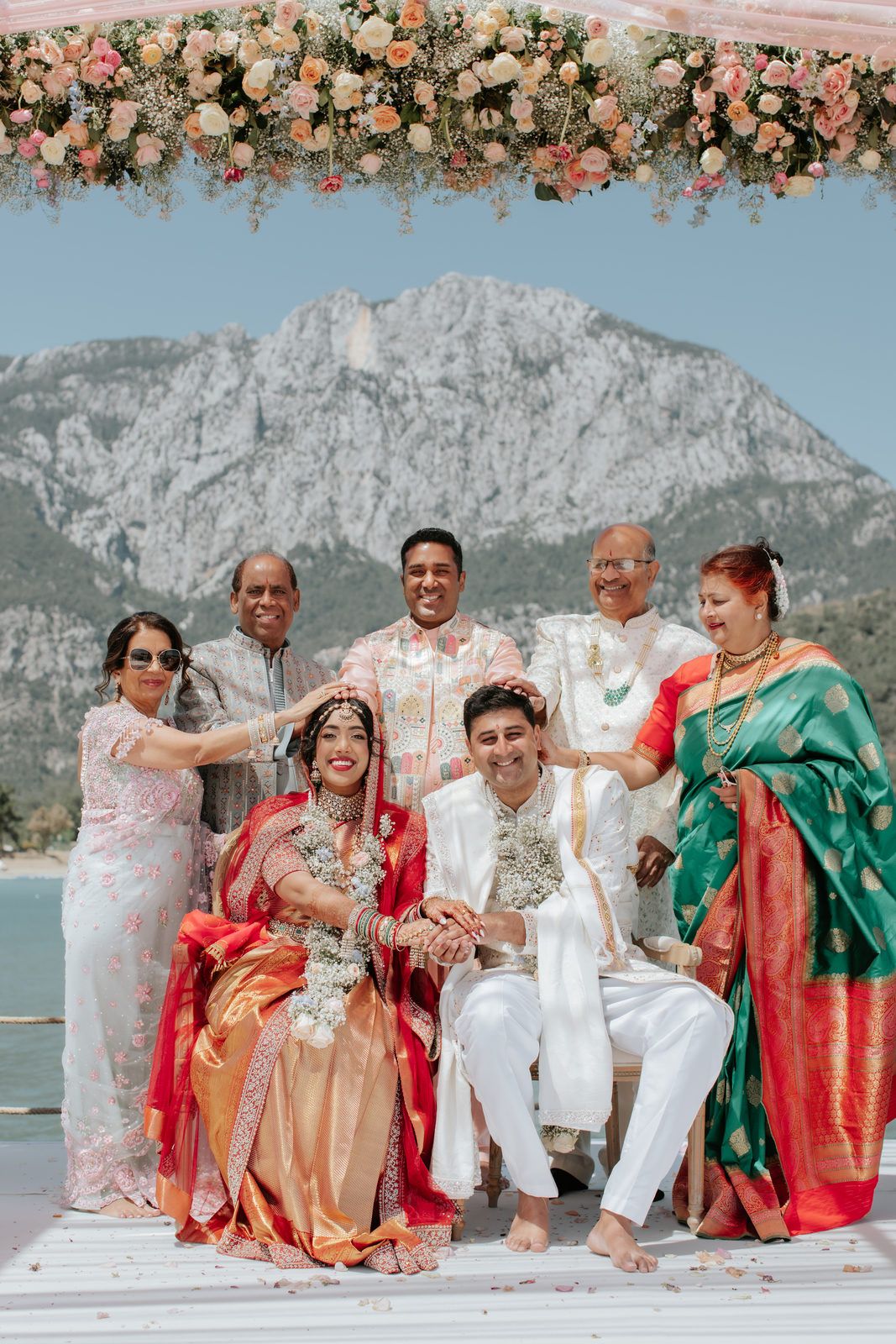 Indian wedding couple in traditional attire seated under floral arch with family, mountain lake backdrop.
