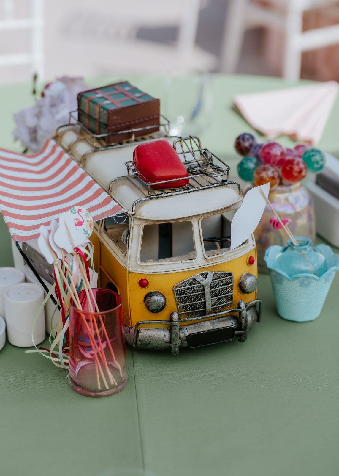 Vintage yellow toy VW bus centerpiece with luggage on roof, surrounded by colorful party decorations on a green table.