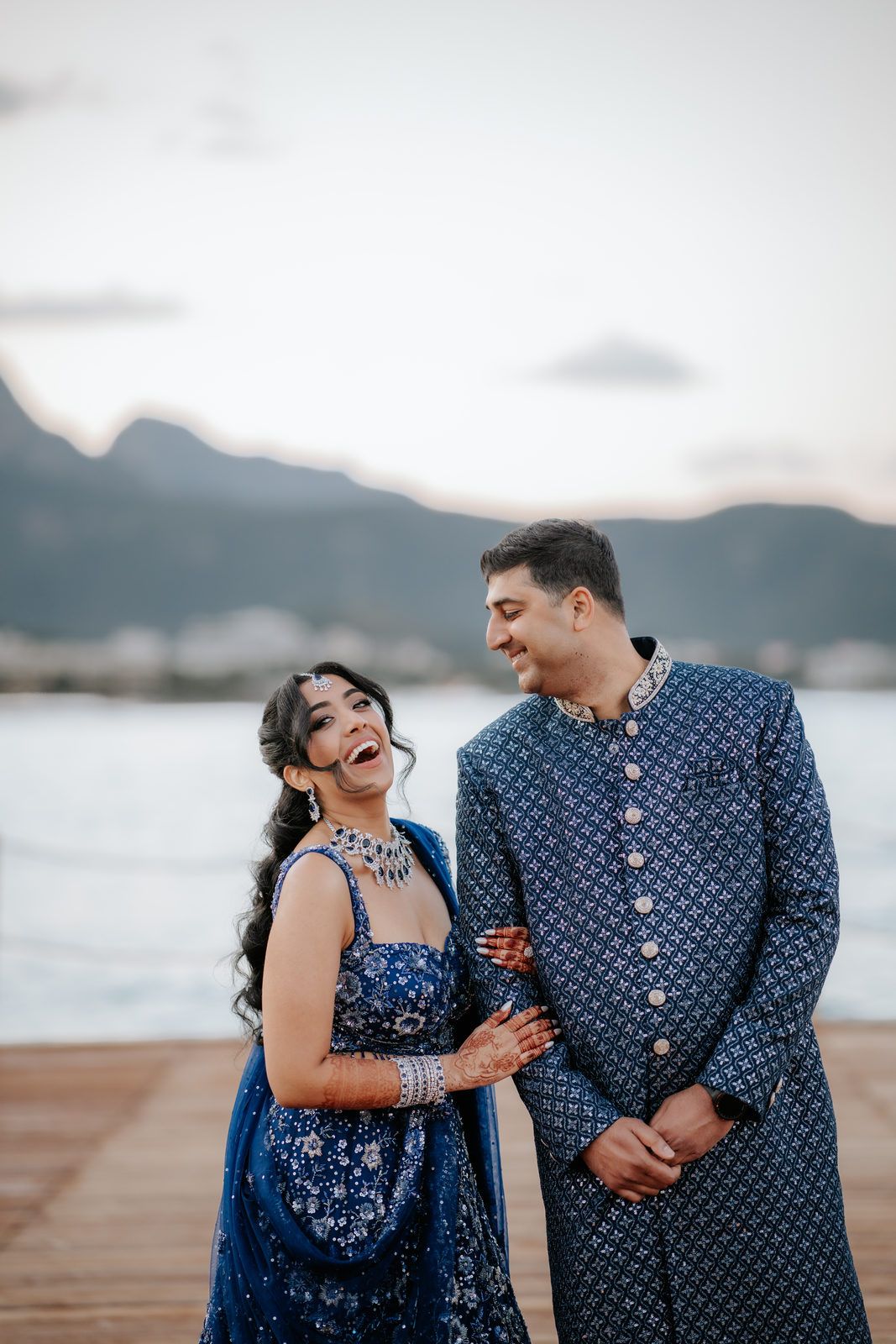 Laughing Indian couple in matching blue traditional wedding outfits on a scenic mountain dock.