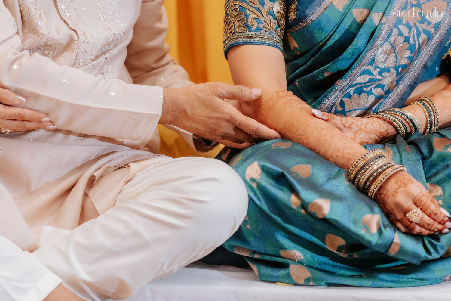 Couple in traditional Indian attire, bride with henna and bangles, groom in white kurta, seated together during ceremony.