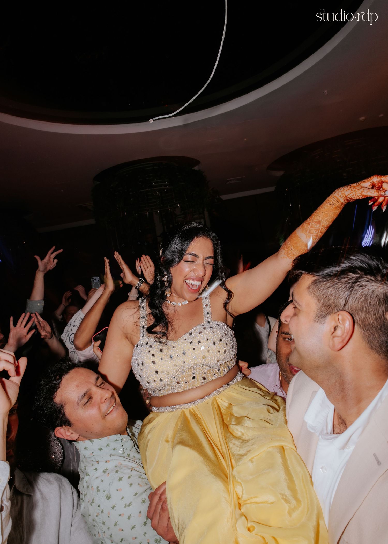 Bride in yellow lehenga laughing joyfully while being lifted by guests on the dance floor at an Indian wedding.